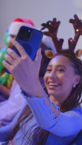 Vertical-Video-Studio-Shot-Of-Gen-Z-Friends-At-Christmas-Sitting-On-Sofa-Wearing-Santa-Hat-And-Reindeer-Antlers-Taking-Selfie-On-Mobile-Phone-1